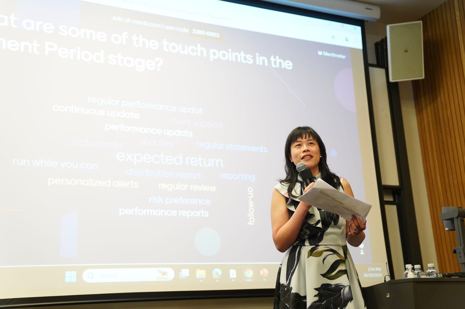 Low-Angle shot of Alice delivering a speech in a lecture hall at UNSW. Behind her, the projector shows a word cloud constructed by the crowd. Alice is wearing a floral dress, smiling as she looks a the crowd.